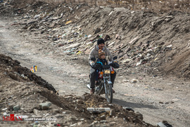 یک موتور سوار با کودک خردسالش در حال عبور از نزدیکی محل جمع آوری زباله های بازیافتی در حومه شهر ری است.